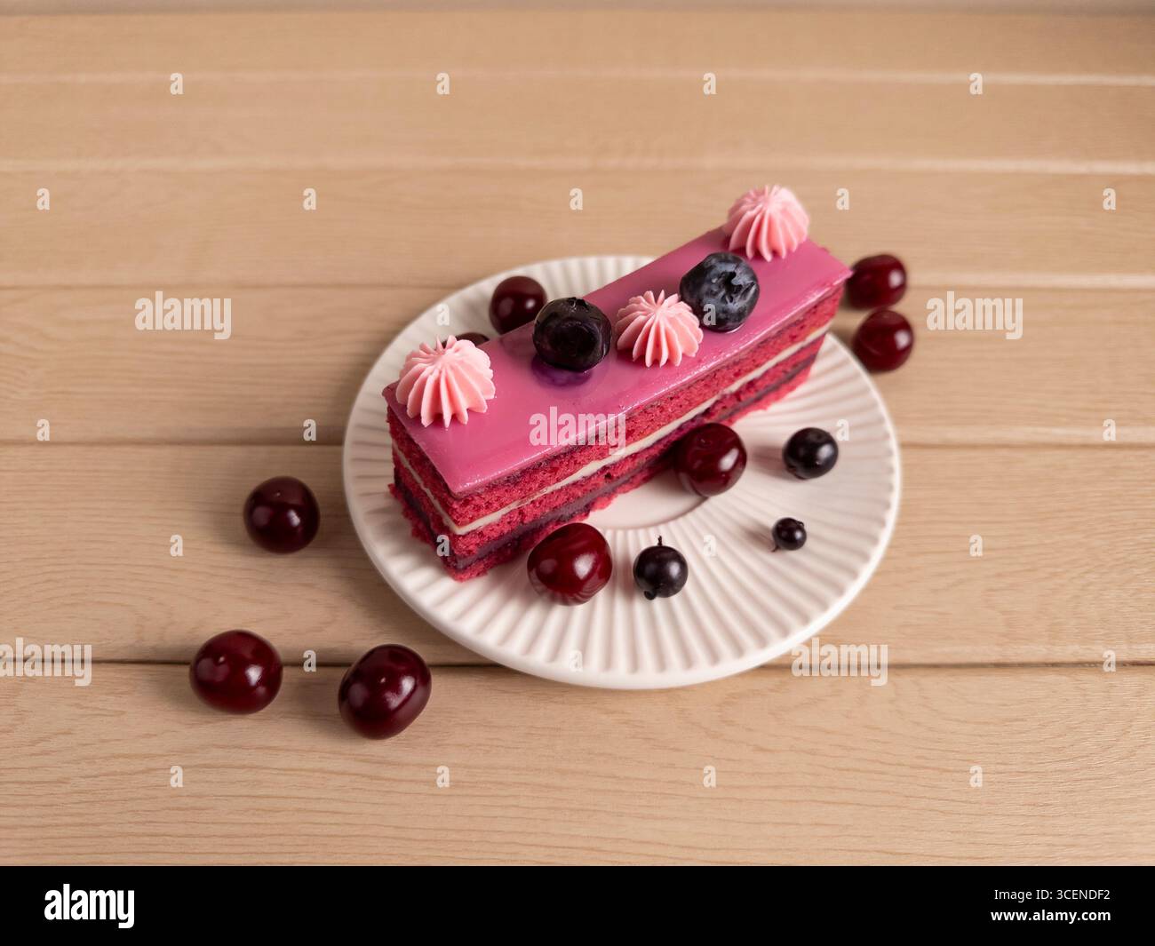 Elegantes Stück Kirsch- und Beerenmousse-Kuchen mit Schichten Sahne und rosa Glasur, garniert mit Heidelbeeren und Kirschen, serviert auf einer weißen Keramik-pla Stockfoto