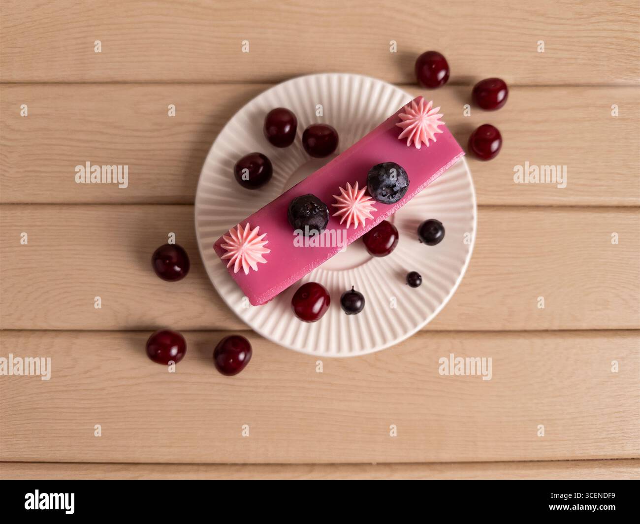 Minimalistischer Blick von oben auf eine rosafarbene Kirsch- und Heidelbeermousse-Tortenscheibe mit Schlagsahne und frischen Beeren, serviert auf einem weißen Teller über einem Stockfoto