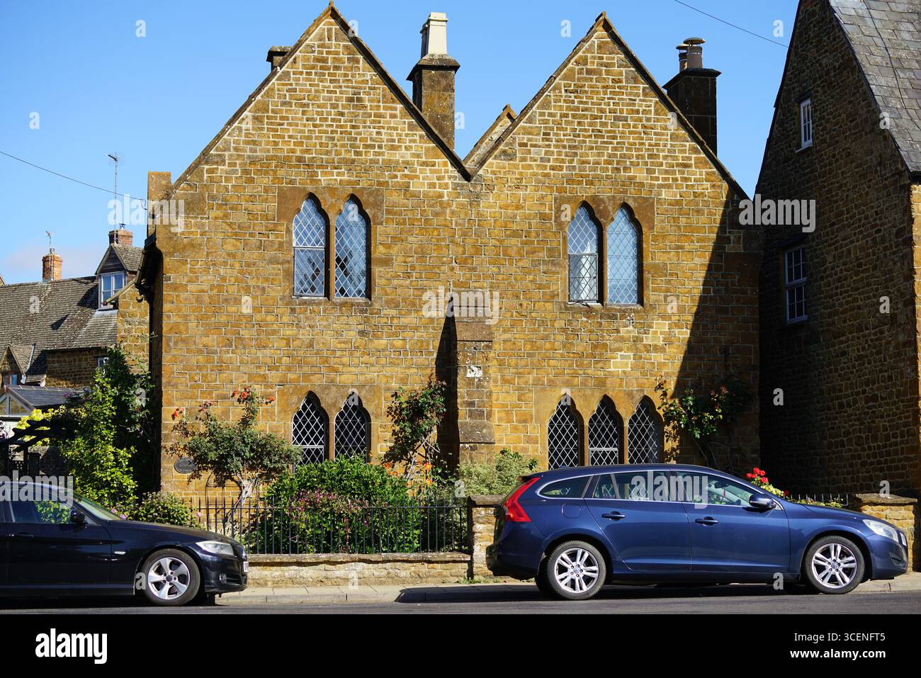 Historisches Steingebäude mit gotischen Fenstern und geparkten Autos an einem sonnigen Tag. Adderbury, England Stockfoto
