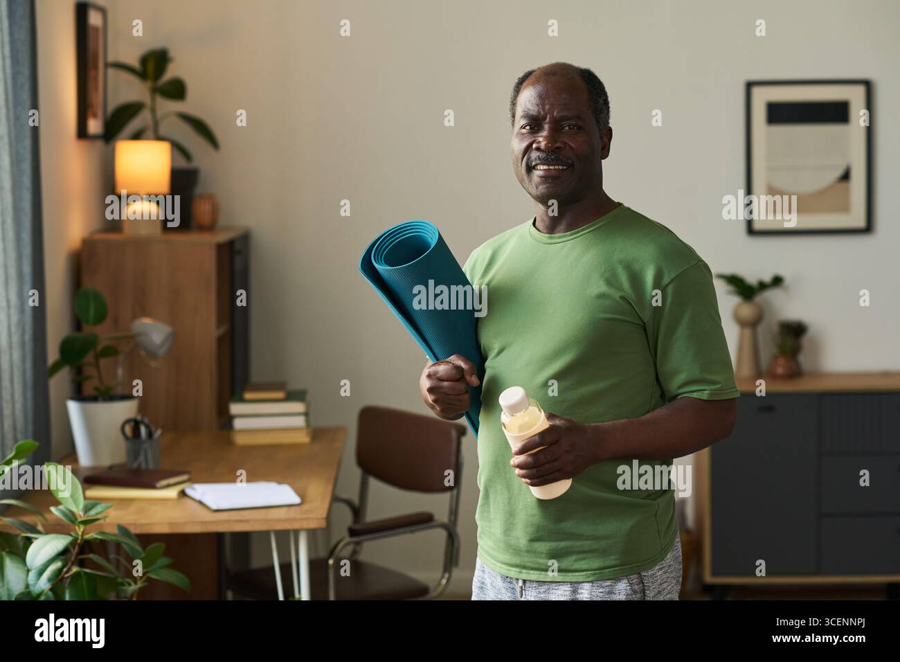 Senior Black man steht im Home Office mit Yogamatte und Wasserflasche, lächelnd und blickend nach vorne, Indoor-Wellness-Konzept mit Pflanzen und gerahmtem Kunstwerk im Hintergrund Stockfoto