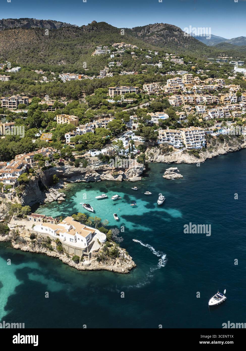 Aus der Vogelperspektive tanzen Boote auf türkisfarbenem Wasser neben der zerklüfteten Küste und den malerischen Gebäuden von Cala Fornells, Illes Balears, Spanien. Stockfoto