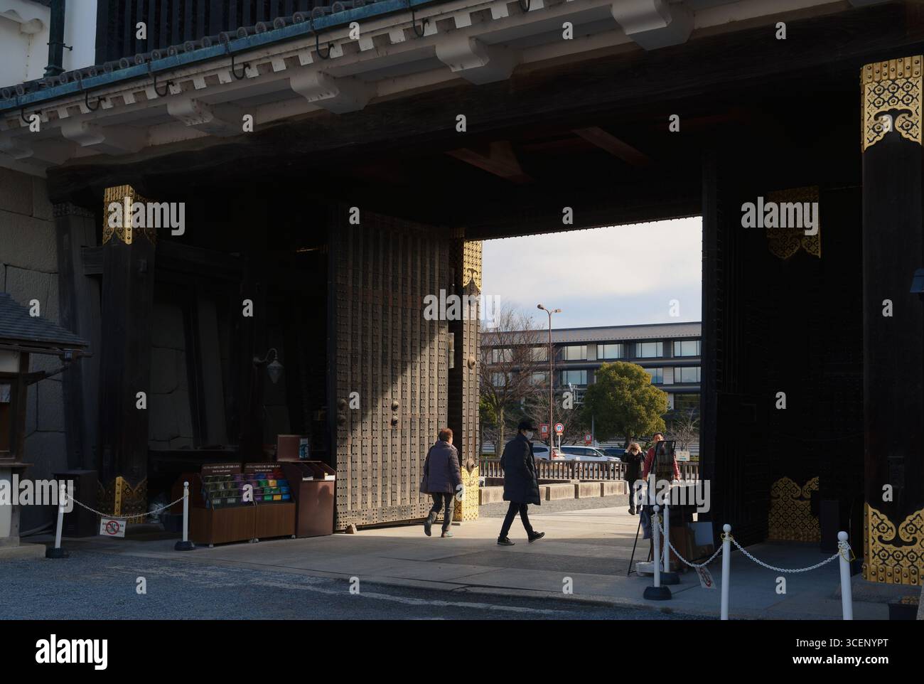Besucher passieren die hölzernen Tore, die mit Golddetails geschmückt sind, auf dem Kaisergelände von Kyoto, und treten vom Schatten in die hellen Winterlichter Stockfoto