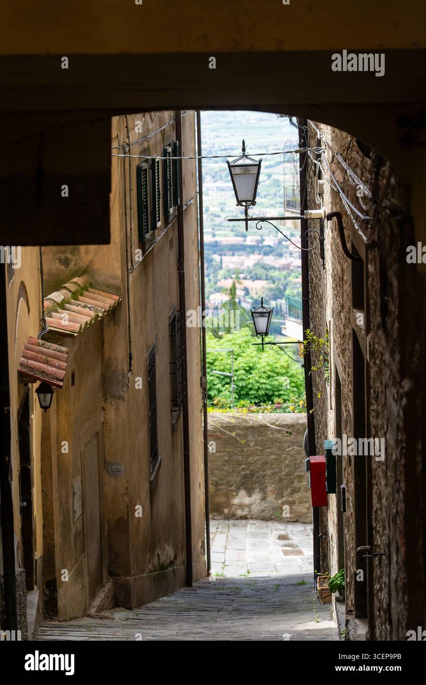Tuscany, Italien - 24. April 2025: Blick auf eine charmante, enge Straße, die von verwitterten Steinhäusern eingerahmt wird, führt zu einem entfernten Panorama der sanften Hügel, die in ein weiches, trübes Licht getaucht sind. Stockfoto