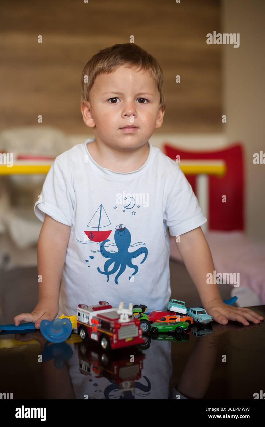 Ein kleines Kind steht mit einem ernsten Ausdruck, während es von bunten Spielzeugautos auf einem Tisch umgeben ist. Die Atmosphäre ist ruhig und kontrastiert seine offensichtliche Dissa Stockfoto
