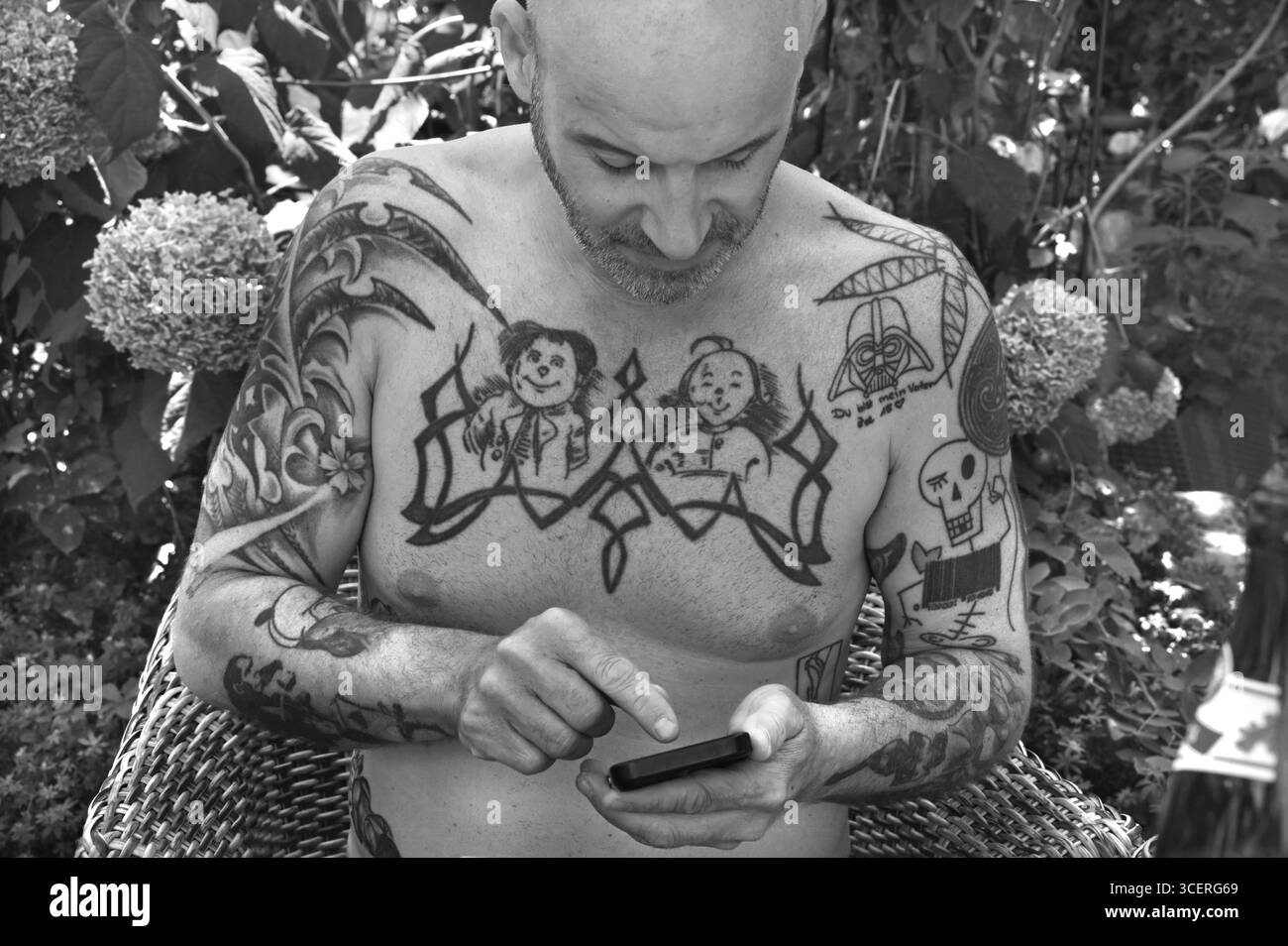 Junger Mann auf Handy, mit Tätowierungen auf Armen und Oberkörper, sitzt unter einem Sonnenschirm im Garten, schwarz-weiß, Baden-Württemberg, Deutschland Stockfoto