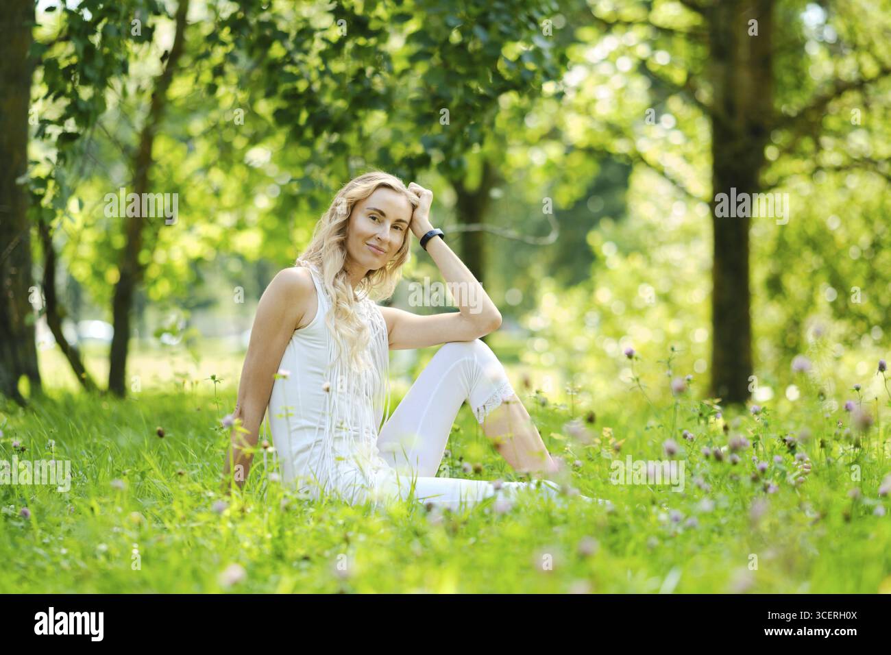 Eine Frau mit langen blonden Haaren sitzt auf weichem Gras, umgeben von blühenden Blumen in einem lebhaften Park. Das Sonnenlicht zieht durch Bäume und schafft ein ruhiges A Stockfoto