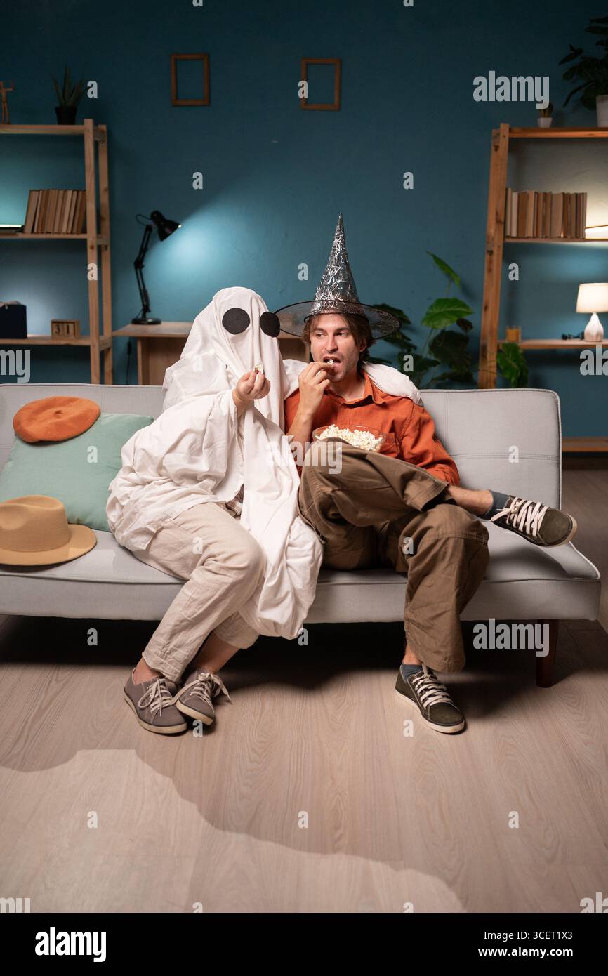 Horrorgespenst in weißem Laken und Mann in Hemd mit Popcorn, der auf dem Sofa sitzt und Filme guckt Stockfoto