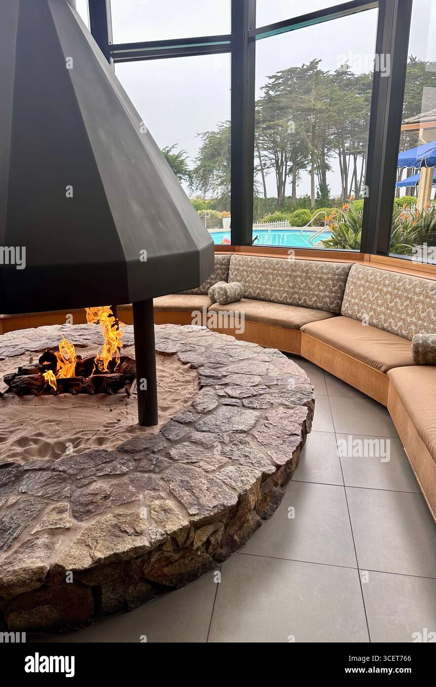Innenraum-Lounge mit Steinfeuer und kreisförmigem Sitzbereich mit Blick auf den Außenpool und die Bäume in einem Resort. - Smartphone-aufgenommenes Stockfoto