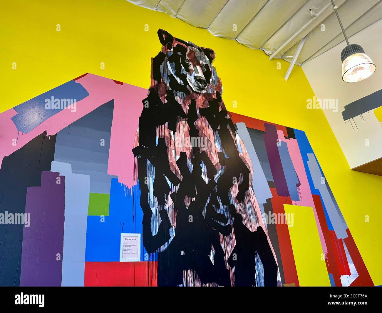 Abstraktes Bärengemälde auf einer hellgelben Wand mit kräftigen geometrischen Formen in Pink, Blau, Rot, Grau und Schwarz. - Smartphone-aufgenommenes Stockfoto