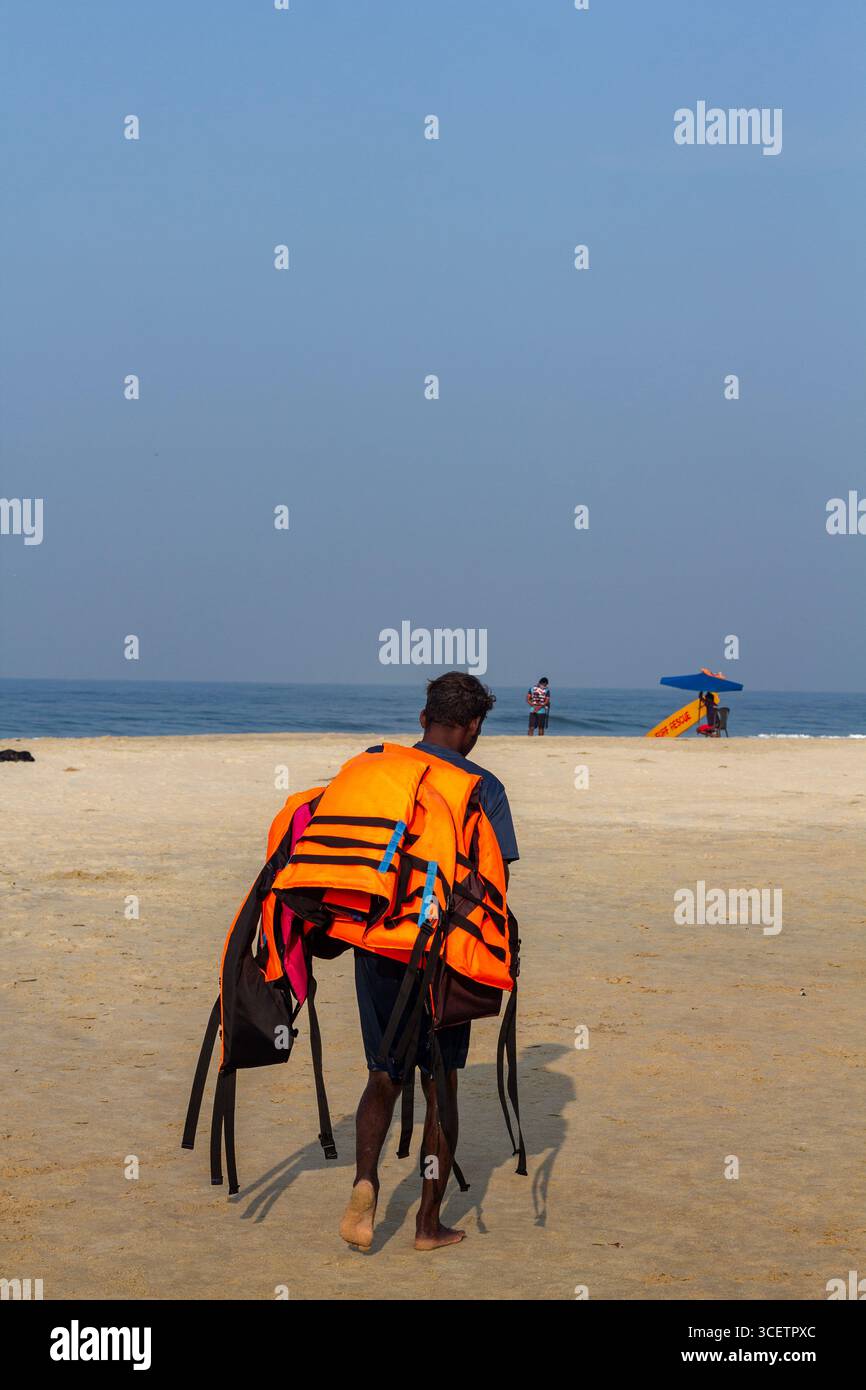 Ein Mann, der mehrere leuchtend orange Schwimmwesten trägt, läuft barfuß an einem Sandstrand in Richtung Meer, mit klarem blauen Himmel und Rettungsschwimmer hinten. Stockfoto