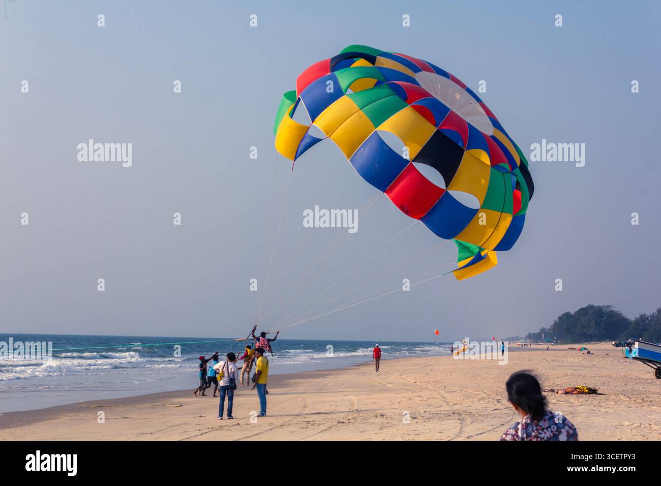 Ein lebendiger, bunter Fallschirm erfüllt den Himmel über einem Sandstrand, wobei an einem hellen, klaren Tag ein Boot und ein Wagen im Hintergrund zu sehen sind Stockfoto