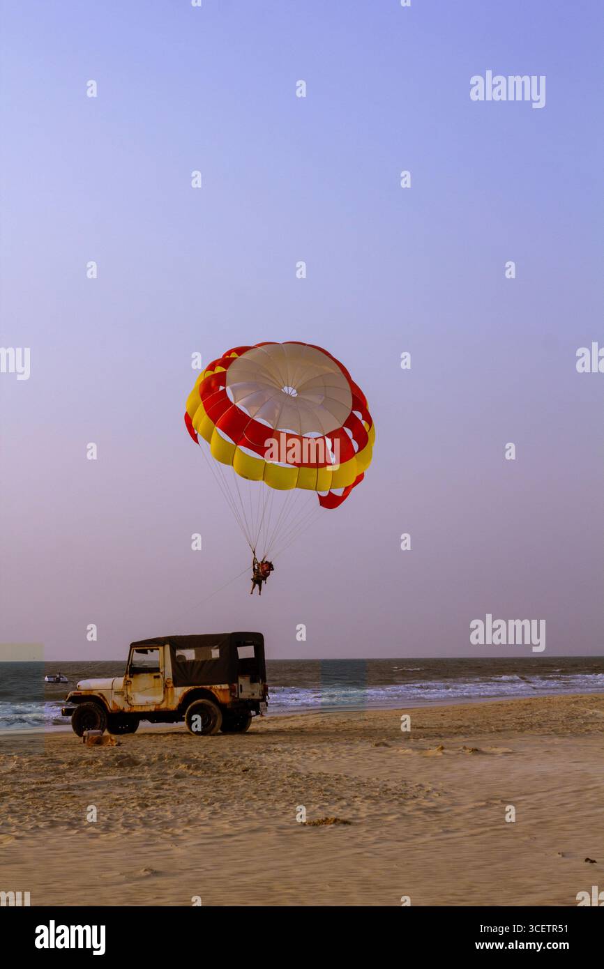 Ein lebendiger, bunter Fallschirm erfüllt den Himmel über einem Sandstrand, wobei an einem hellen, klaren Tag ein Boot und ein Wagen im Hintergrund zu sehen sind Stockfoto