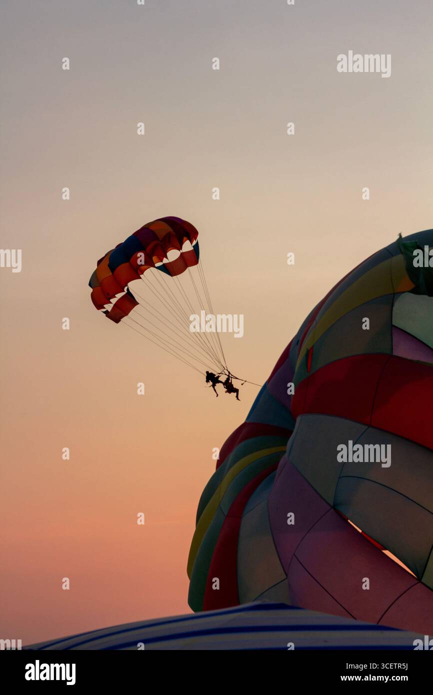 Ein lebendiger, bunter Fallschirm erfüllt den Himmel über einem Sandstrand, wobei an einem hellen, klaren Tag ein Boot und ein Wagen im Hintergrund zu sehen sind Stockfoto