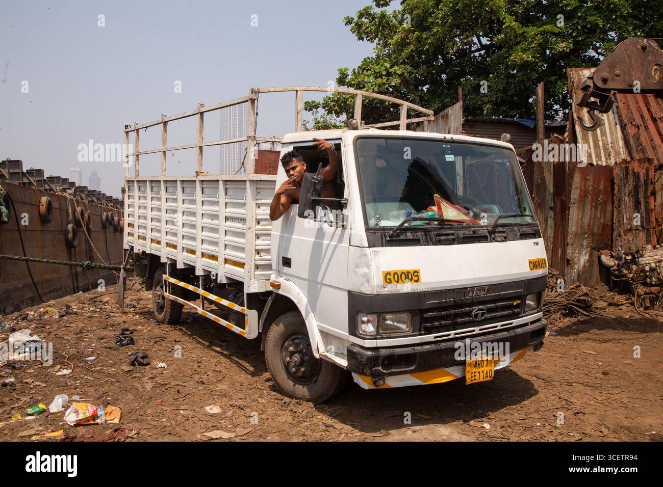 Ein Lkw für den Transport von weißen Gütern steht auf einem staubigen Industriegelände, umgeben von Altartikeln und Metallschuppen, unter Tageslicht mit Bäumen Stockfoto