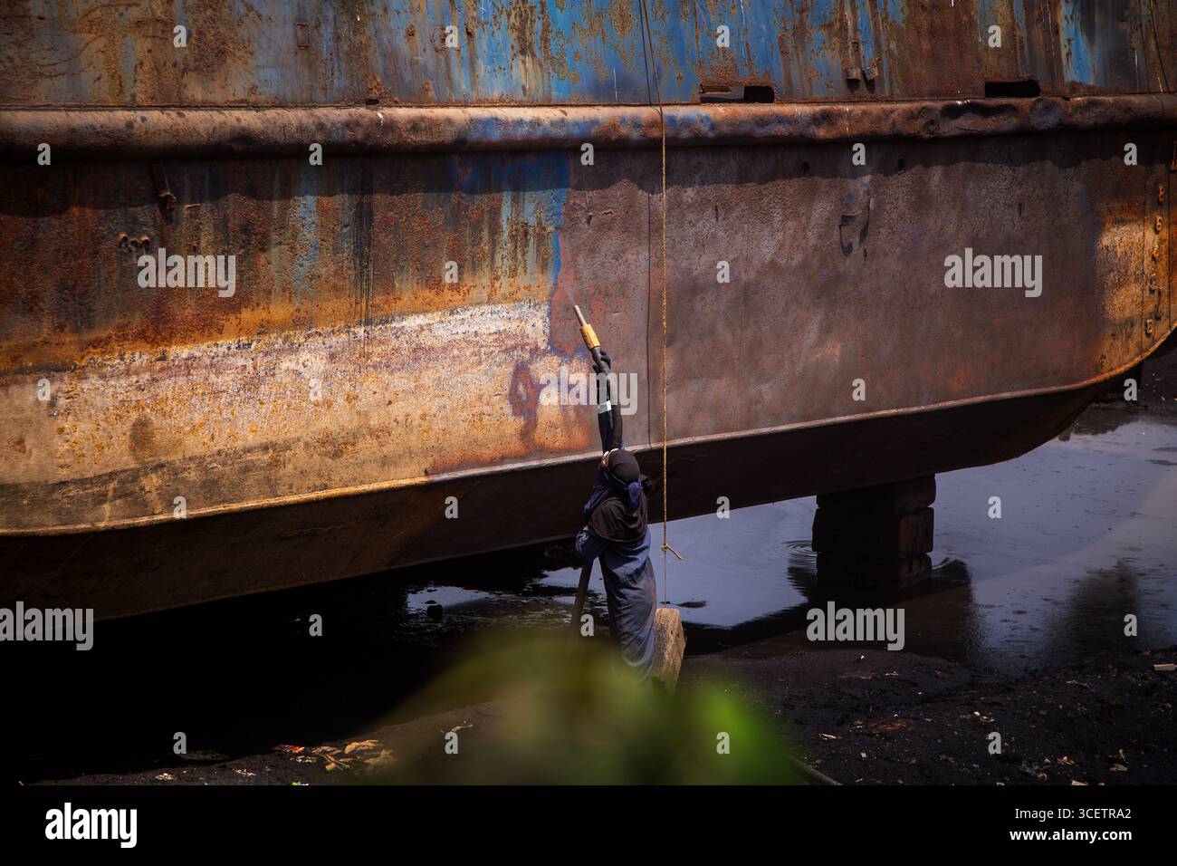 Dieses Bild zeigt einen Arbeiter, der den großen, verrosteten Rumpf eines Schiffes mit einem Industriespray oder Hochdruckreiniger reinigt oder lackiert. Stockfoto