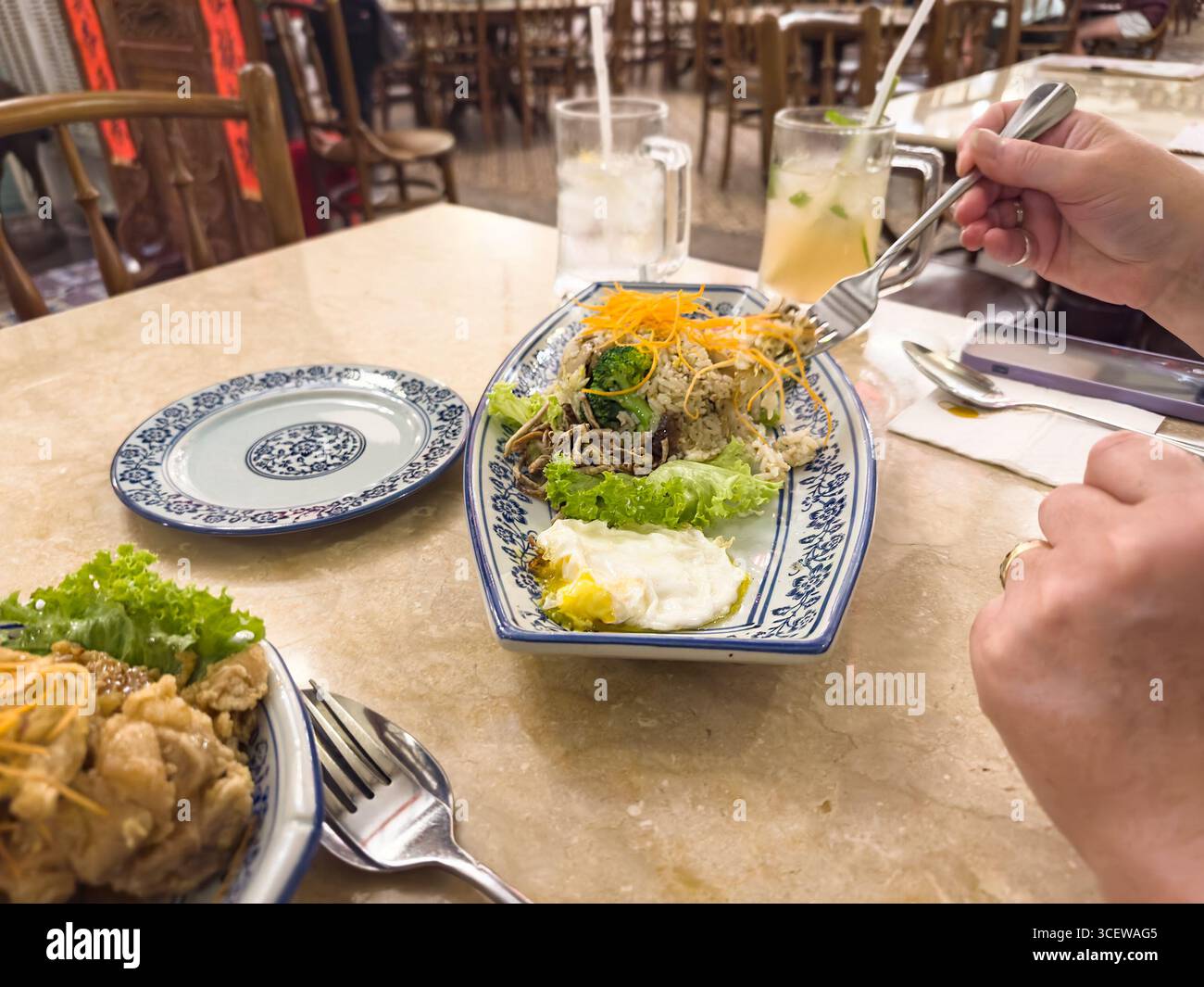 Person, die am asiatischen Restauranttisch mit einem Nudelgericht Eiergemüse auf traditionellem chinesischem Geschirr und geeistem Zitronentee Malacca malaysia speisen kann - Smartphone-aufgenommenes Stockfoto