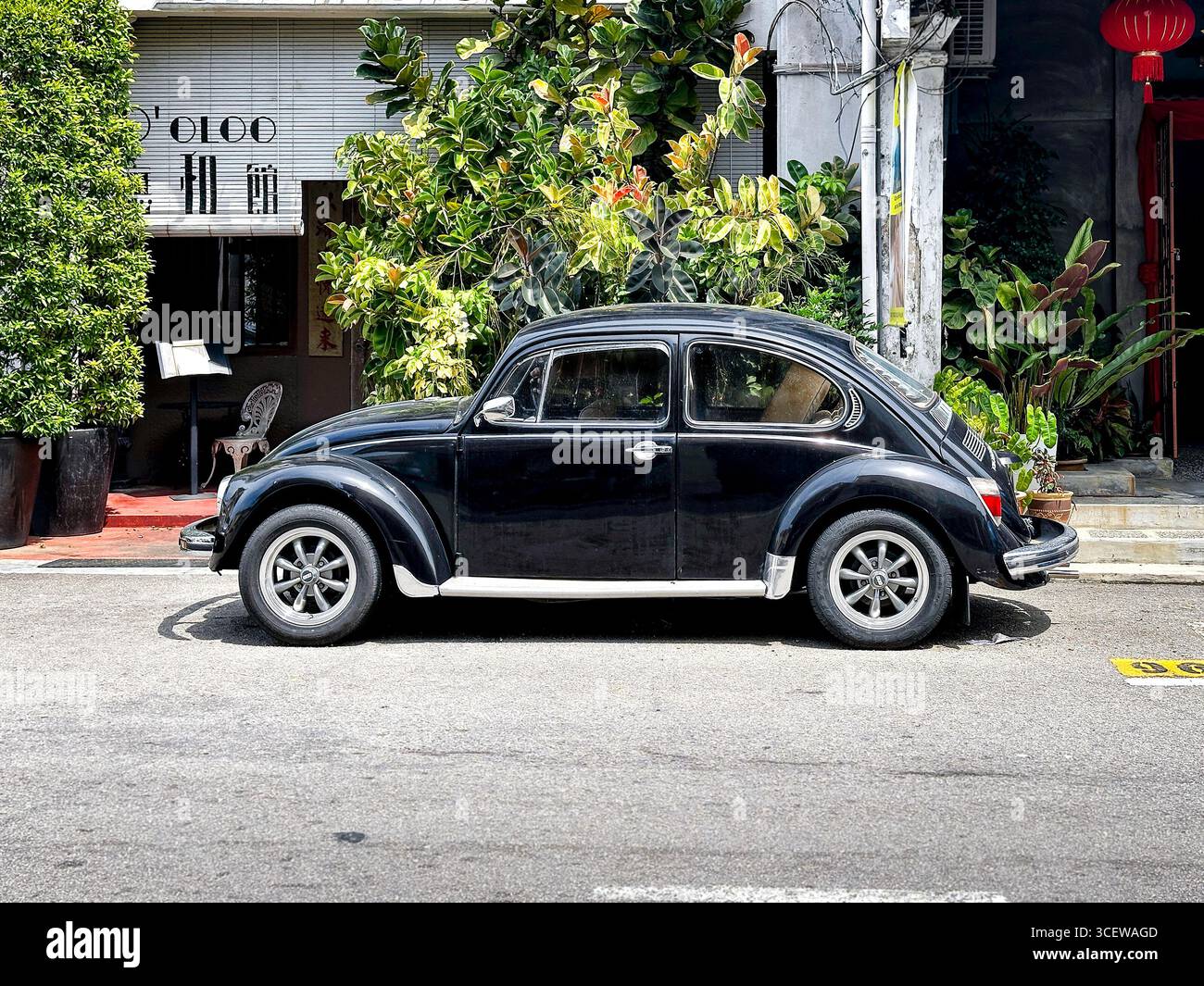 Seitenansicht des schwarzen Vintage Volkswagen Käfers Typ 1 auf der Straße mit üppigen grünen Pflanzen und Gebäudeeingang im Hintergrund Malacca malaysia Stockfoto