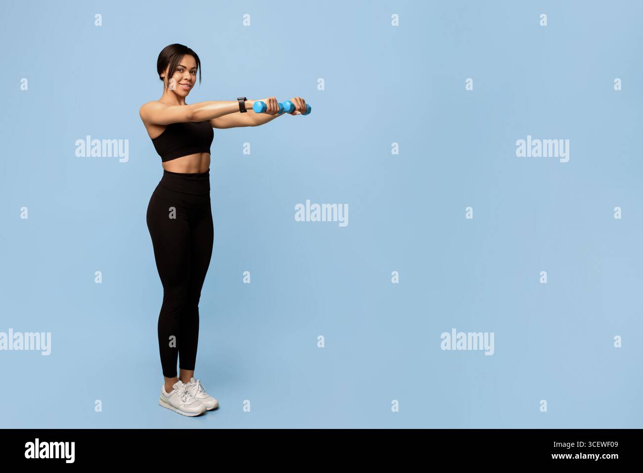 Armtraining. Glückliche junge afroamerikanische Sportlerin, die mit zwei Kurzhanteln auf blauem Hintergrund trainiert Stockfoto