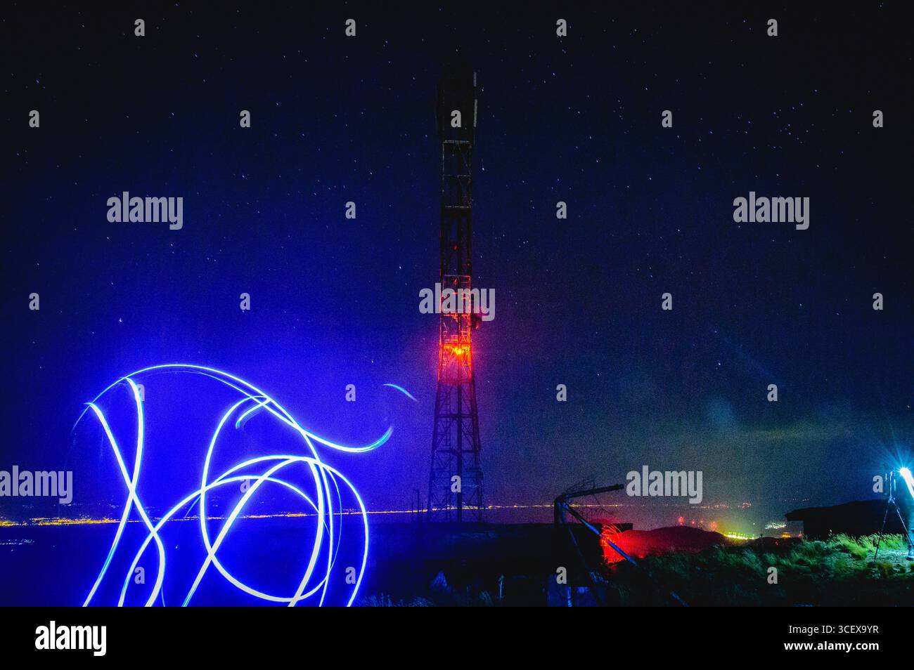 Kreative Langzeitbelichtung von Lichtspuren vor einem rot beleuchteten Telekommunikationsturm auf einem Hügel, unter einem klaren Sternenhimmel und glühenden Lichtern der Stadt. Stockfoto