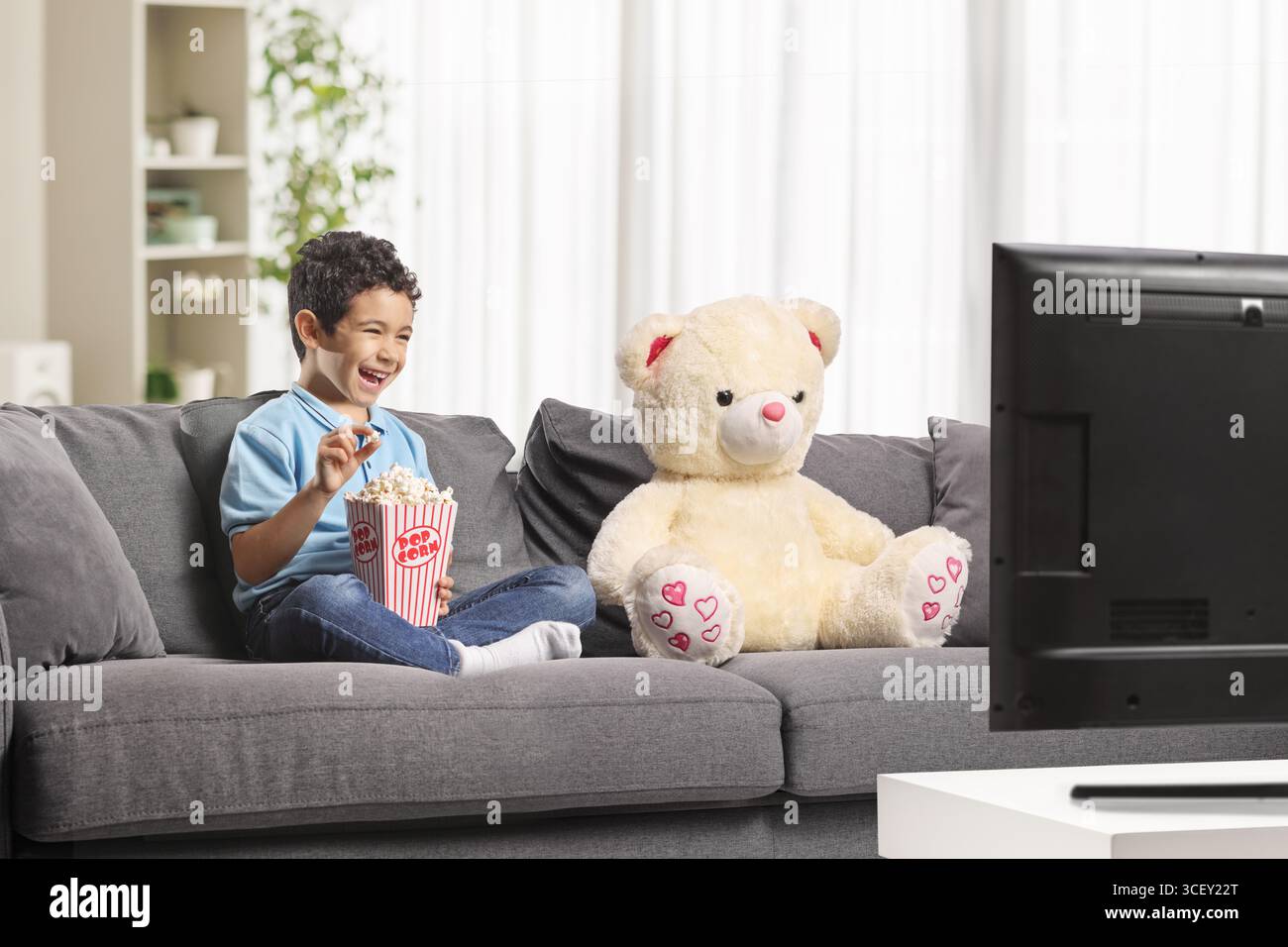 Junge isst Popcorn und sieht mit seinem Teddybären fern Stockfoto