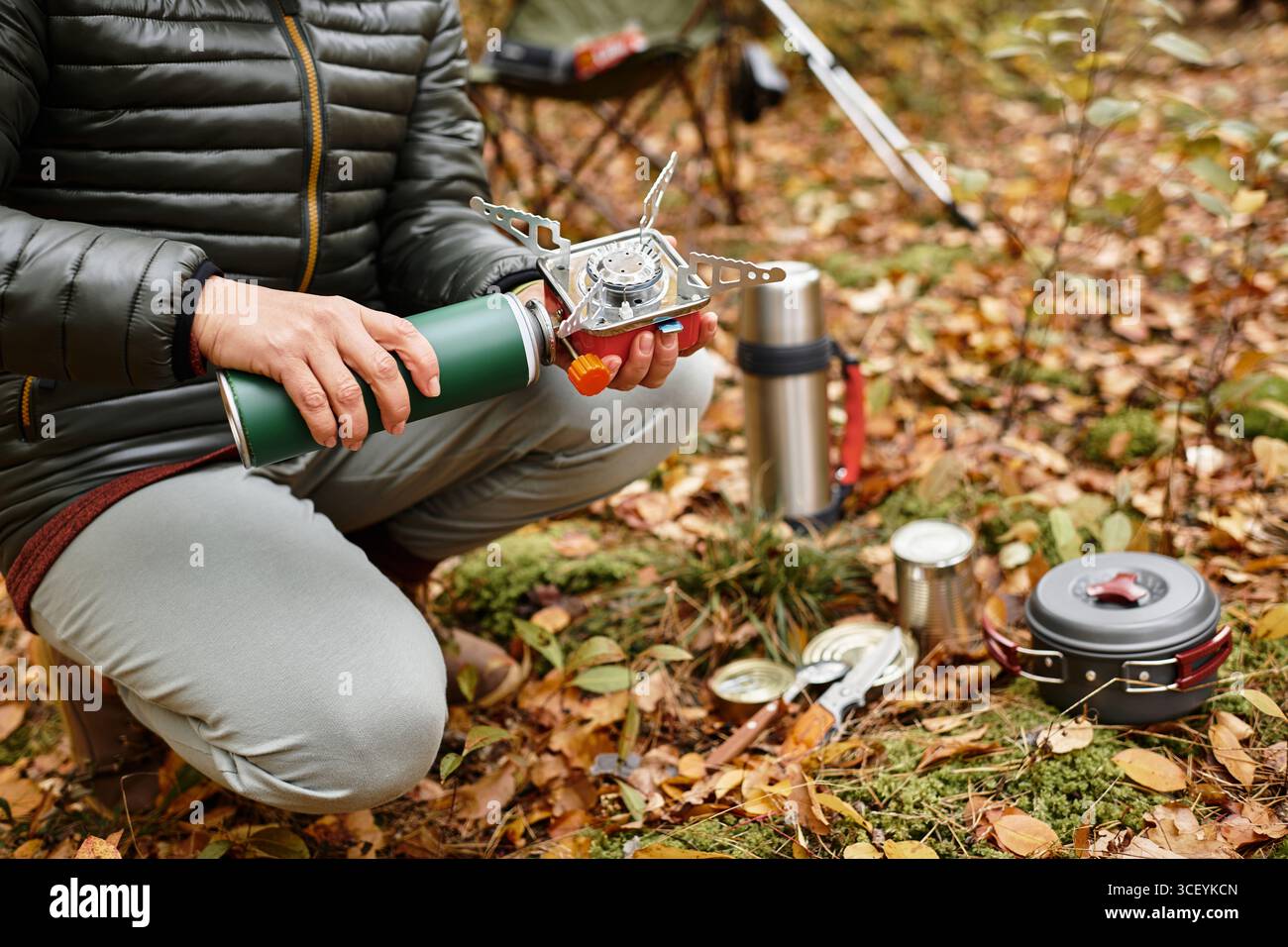 Eine ältere Frau kniet im Herbstlaub und stellt einen tragbaren Herd auf, um eine Mahlzeit im Wald zuzubereiten. Stockfoto