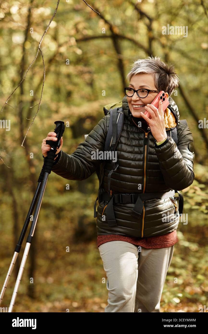 Eine Seniorin durchquert einen üppigen Waldweg, lächelt und redet aufgeregt auf ihrem Telefon. Stockfoto