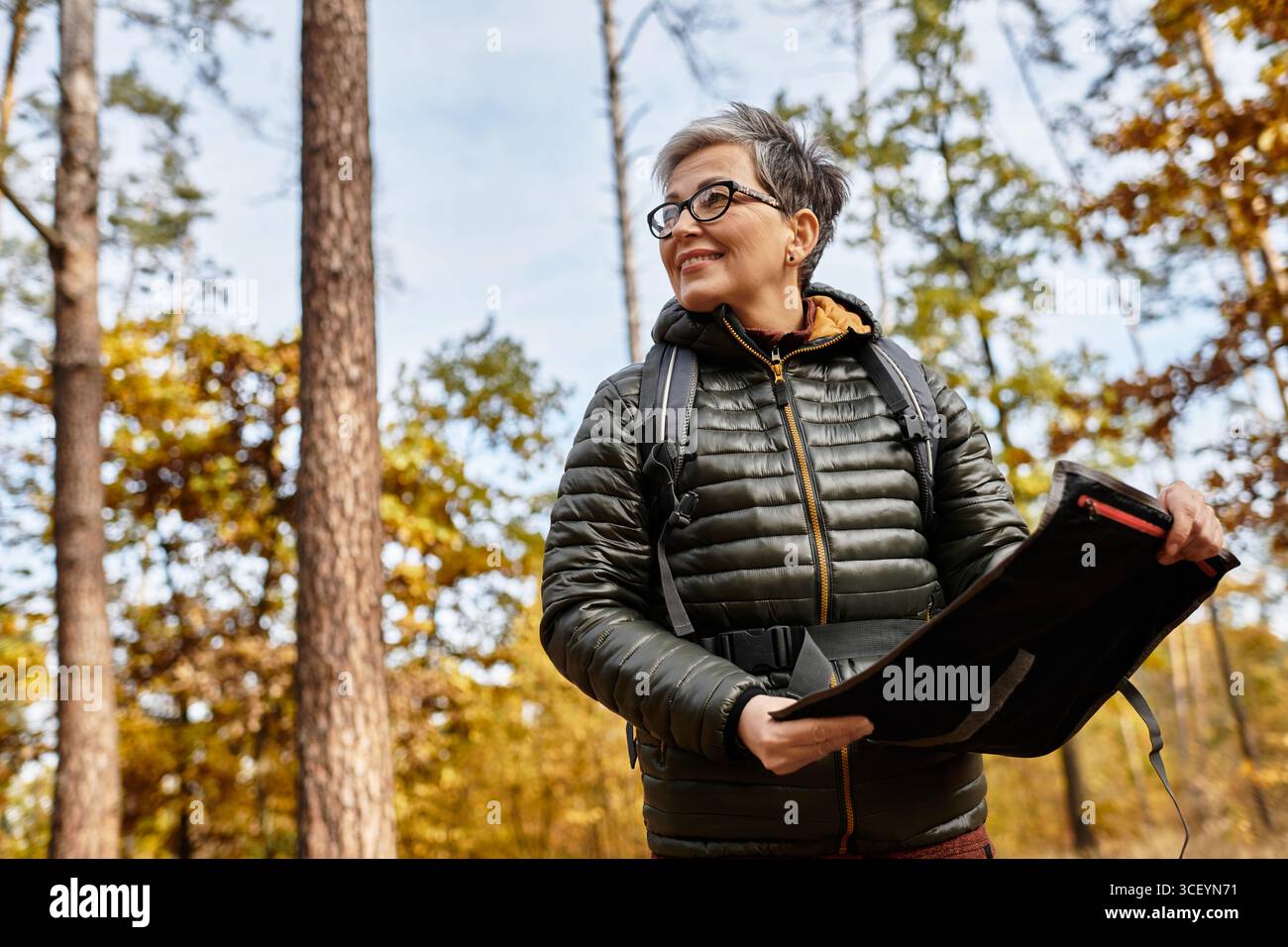 Eine ältere Frau genießt ihre Wanderung im Wald, umgeben von farbenfrohen Herbstlaub und Sonnenschein. Stockfoto