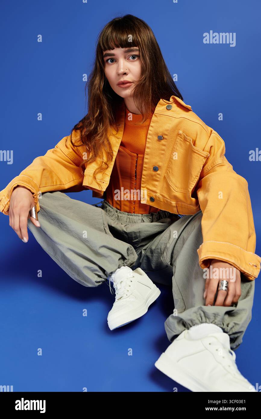Die junge Frau sitzt selbstbewusst und zeigt ihre modische Kleidung und ihren einzigartigen Stil gegen Blau. Stockfoto