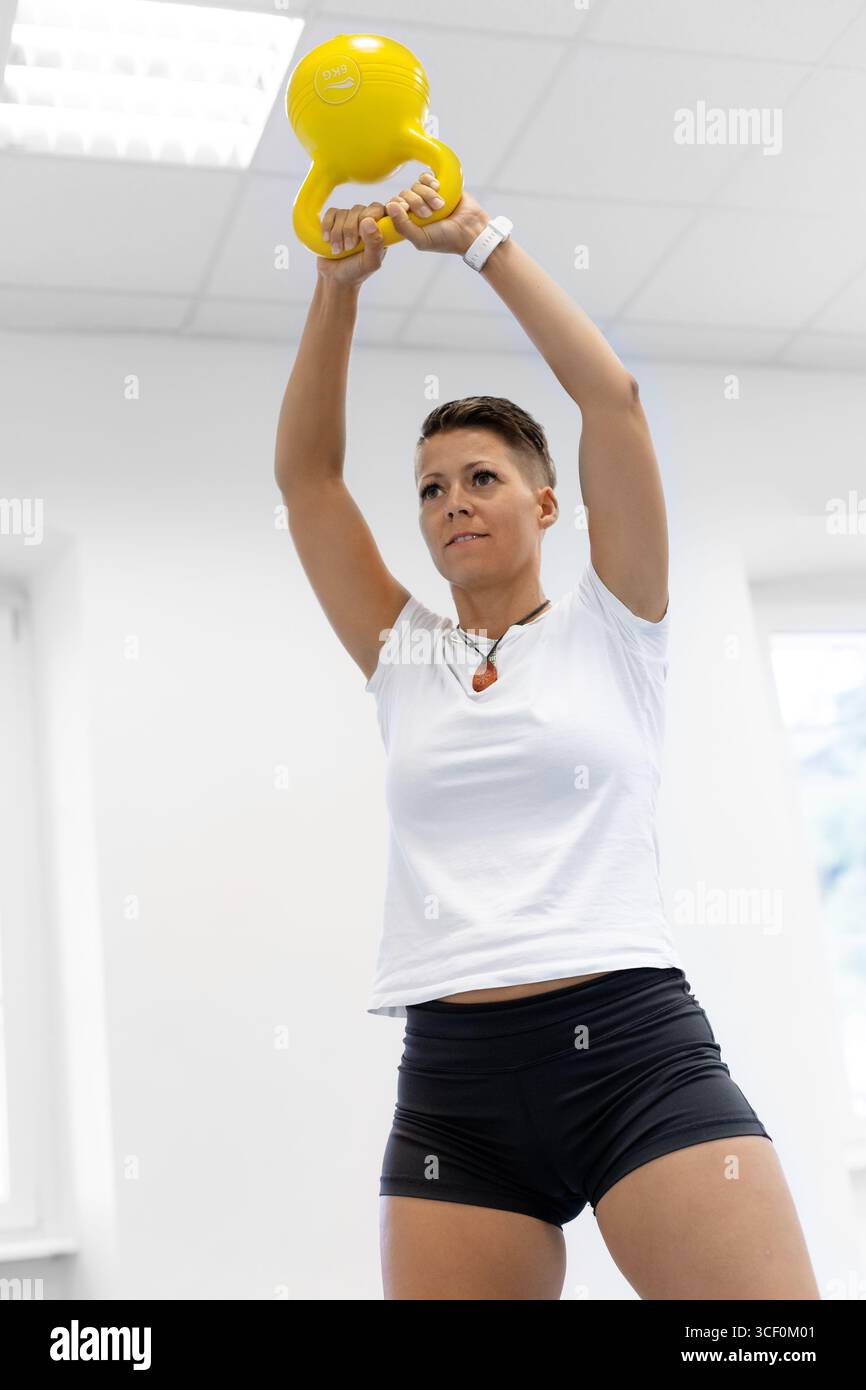 Sportliche Frau, die im Fitnessstudio mit der gelben Kettlebell trainiert und das Gewicht über den Kopf hebt, um Kraft, Ausdauer und Fitness aufzubauen. Stockfoto