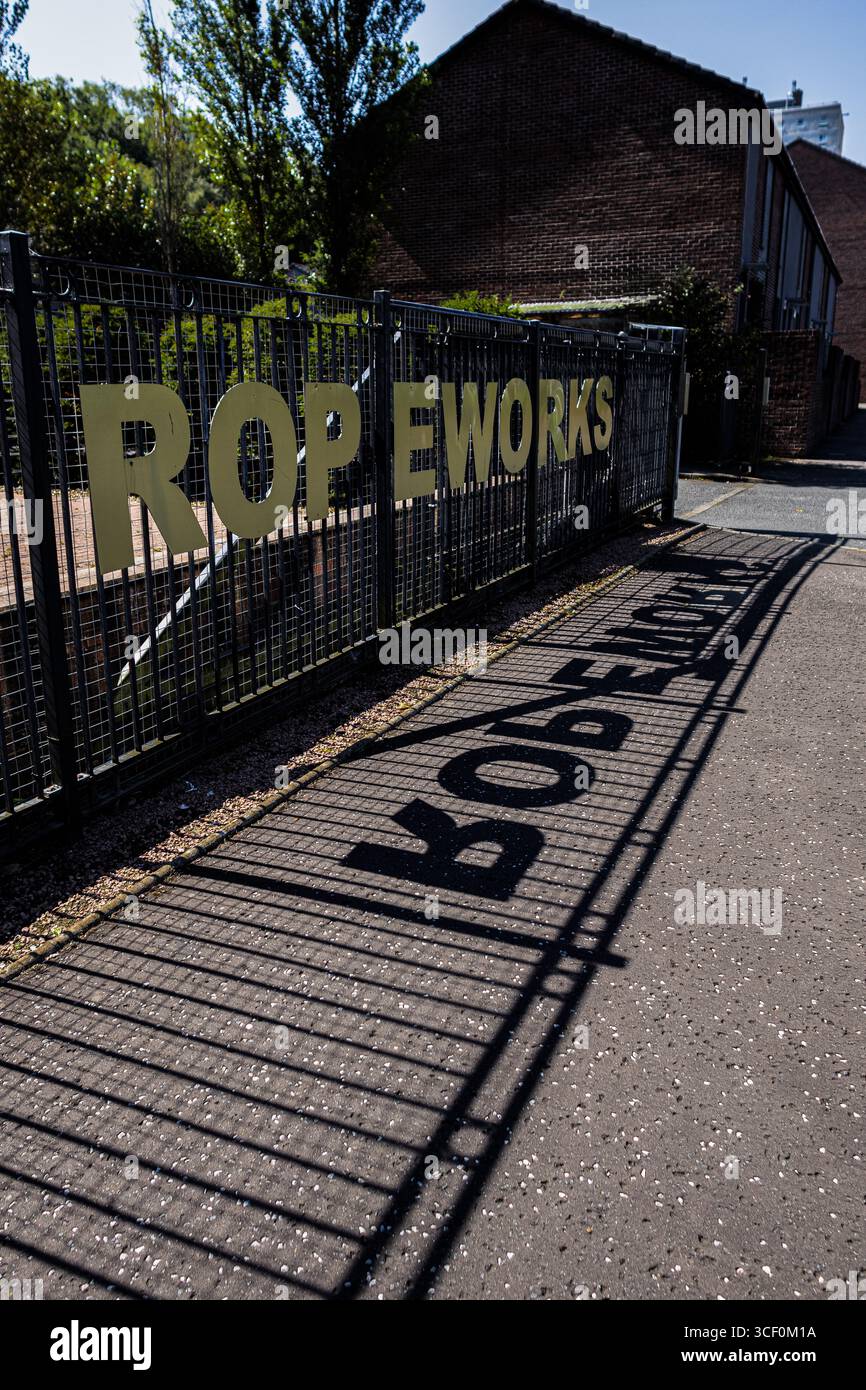 Eine perspektivische Aufnahme eines Zauns mit „ROPEWORKS“ drauf, die einen langen Schatten wirft Stockfoto