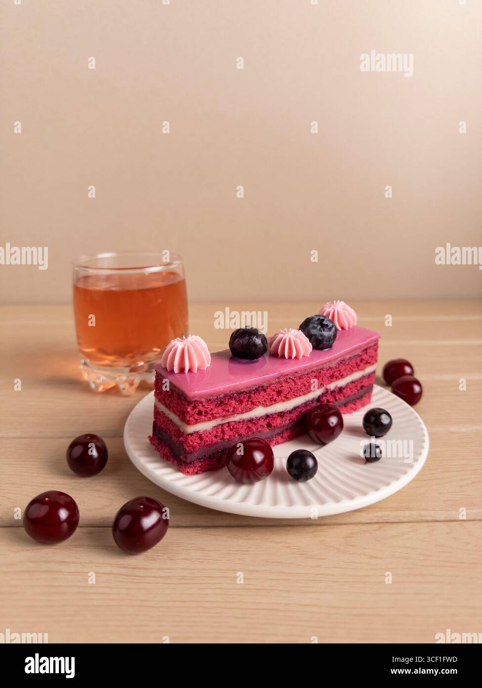 Ein Stück Kuchen mit rosa Zuckerguss, garniert mit Heidelbeeren und Kirschen, begleitet von einem Glas Tee. Das Dessert ist auf einem weißen Stück angeordnet Stockfoto