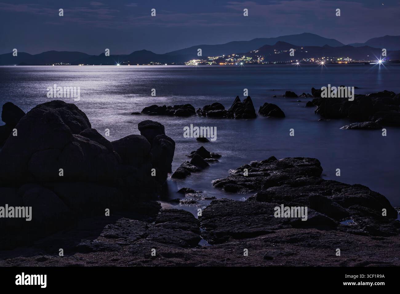 Ruhige Mondszene am Strand des Capo Ferrato in sardinien, italien, mit Felsen, ruhigem Meer und fernen Lichtern der Stadt bei Nacht Stockfoto