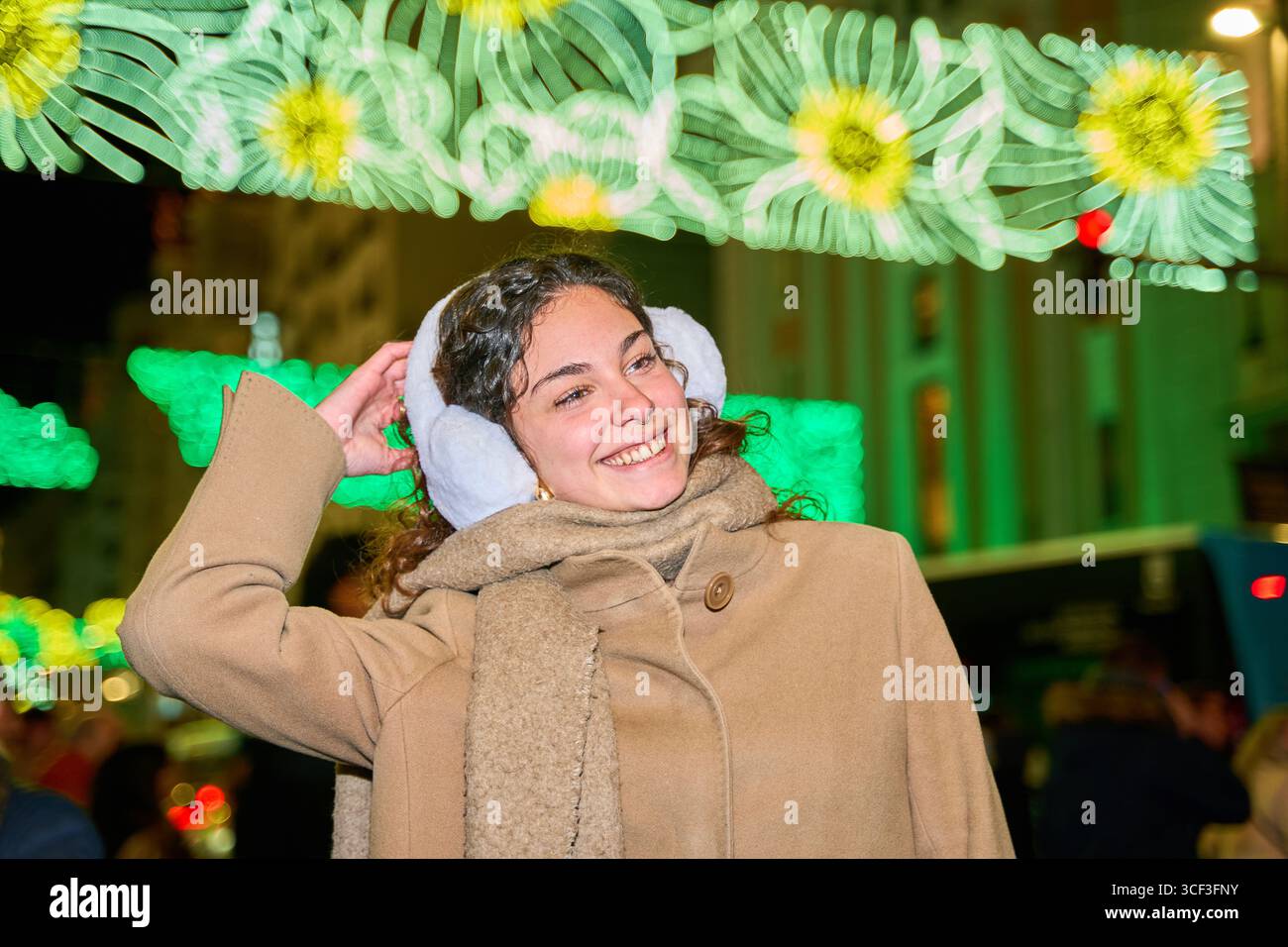 Glückliche junge Frau passt ihre Ohrenschützer an und genießt den weihnachtsgeist unter dekorativen Lichtern Stockfoto
