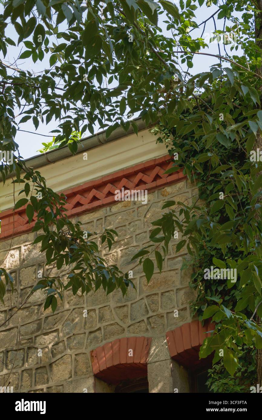 Eine Steinmauer ist mit detaillierten Ziegelsteindekorationen unter hellem Himmel umgeben, umgeben von üppig grünen Blättern, die eine ruhige und einladende Atmosphäre in einem schaffen Stockfoto