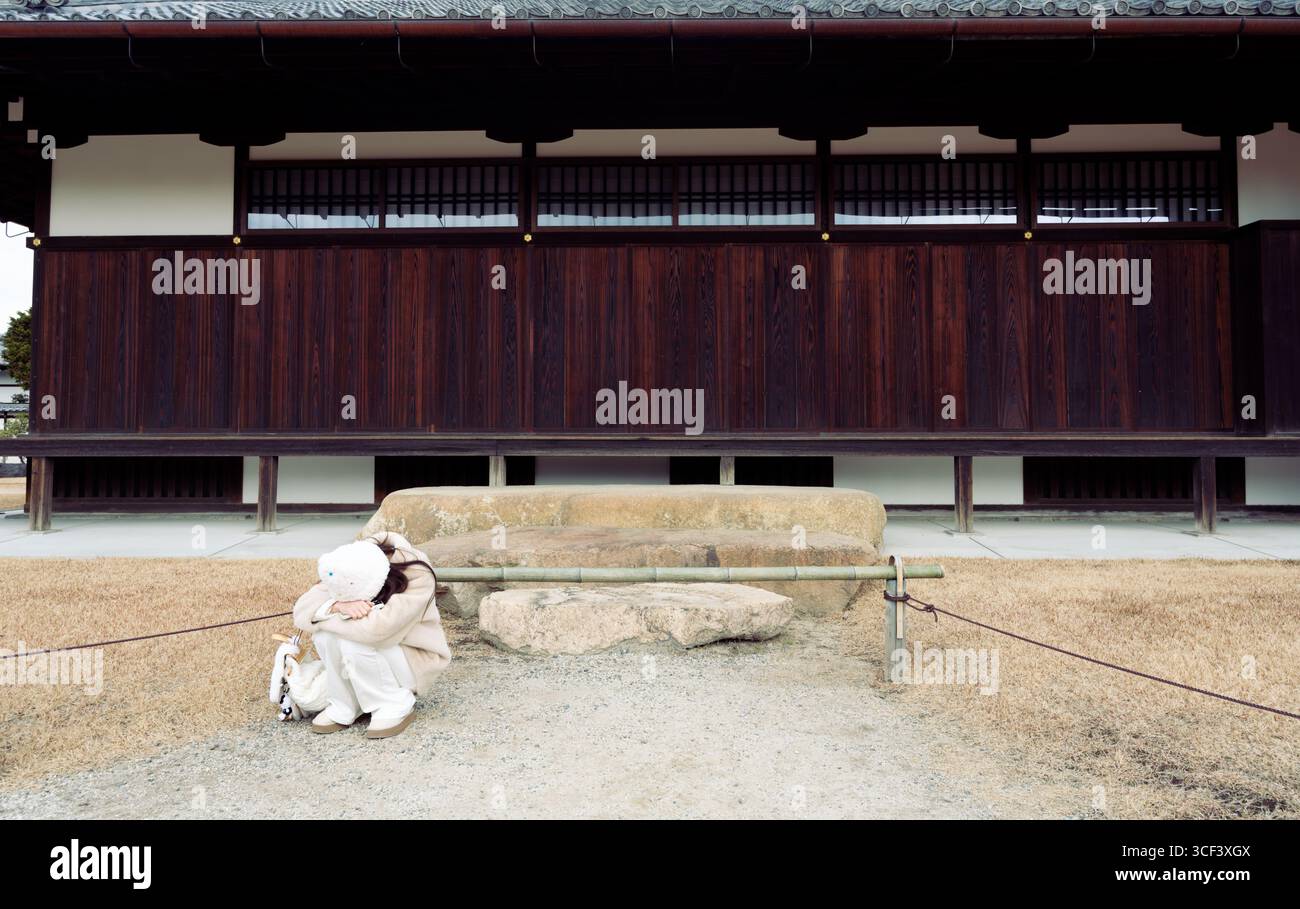 Eine junge Frau in weiß sitzt in ruhiger Traurigkeit eingerollt vor den hölzernen Hallen von Nijo Castle Stockfoto