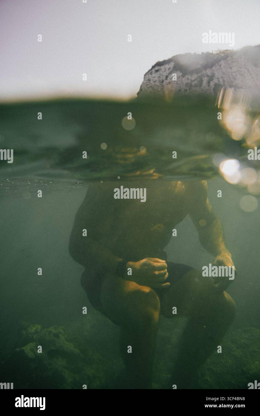 Ein Mann sitzt im Wasser mit den Händen auf den Knien. Das Wasser ist grün und trüb Stockfoto