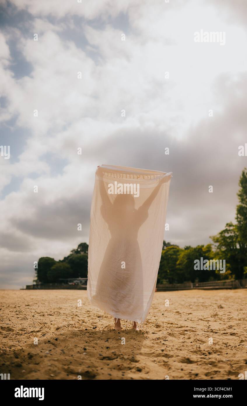 Eine Frau steht an einem Strand mit einer weißen Decke über ihr. Die Decke ist groß genug, um ihren ganzen Körper zu bedecken, und sie hält sie hoch Stockfoto