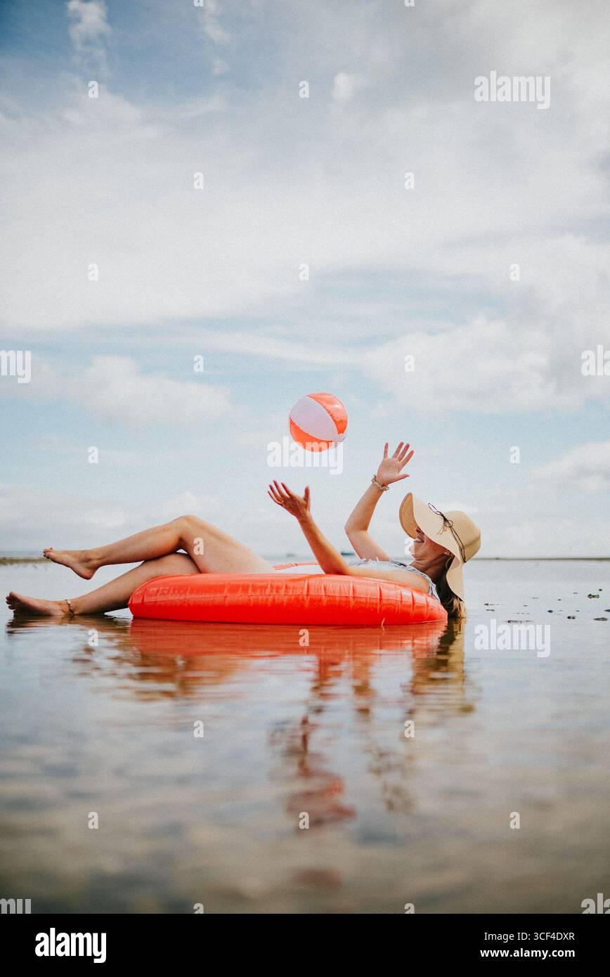 Eine Frau schwimmt in einem roten Schlauchboot mit einem Strandball in der Luft. Die Szene ist entspannt und sorglos, die Frau genießt ihre Zeit im Stockfoto