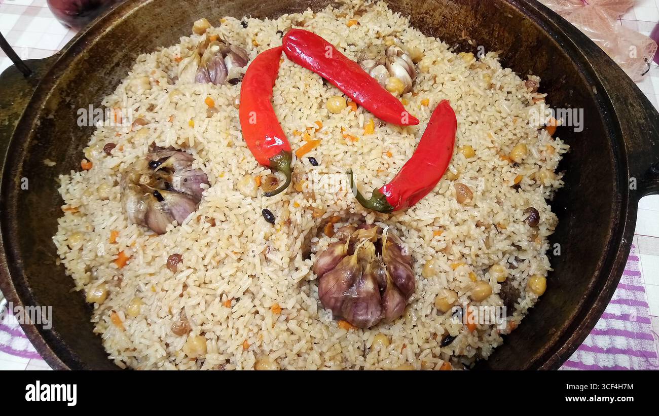 Die Paprika sind kreuzförmig angeordnet. Die Schüssel steht auf einem Tisch. Auf dem Tisch steht ein Kessel mit fertigem Pilaw und rotem Pfeffer. Stockfoto