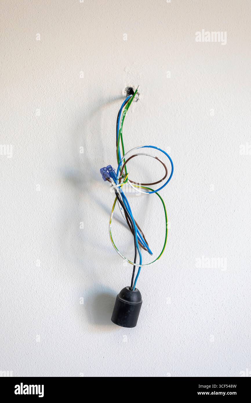 Bunte elektrische Leitungen, die aus einem Loch in einer weißen Wand mit einem Lampenhalter kommen, deuten auf laufende Arbeiten bei Renovierungen oder Neubauten hin Stockfoto