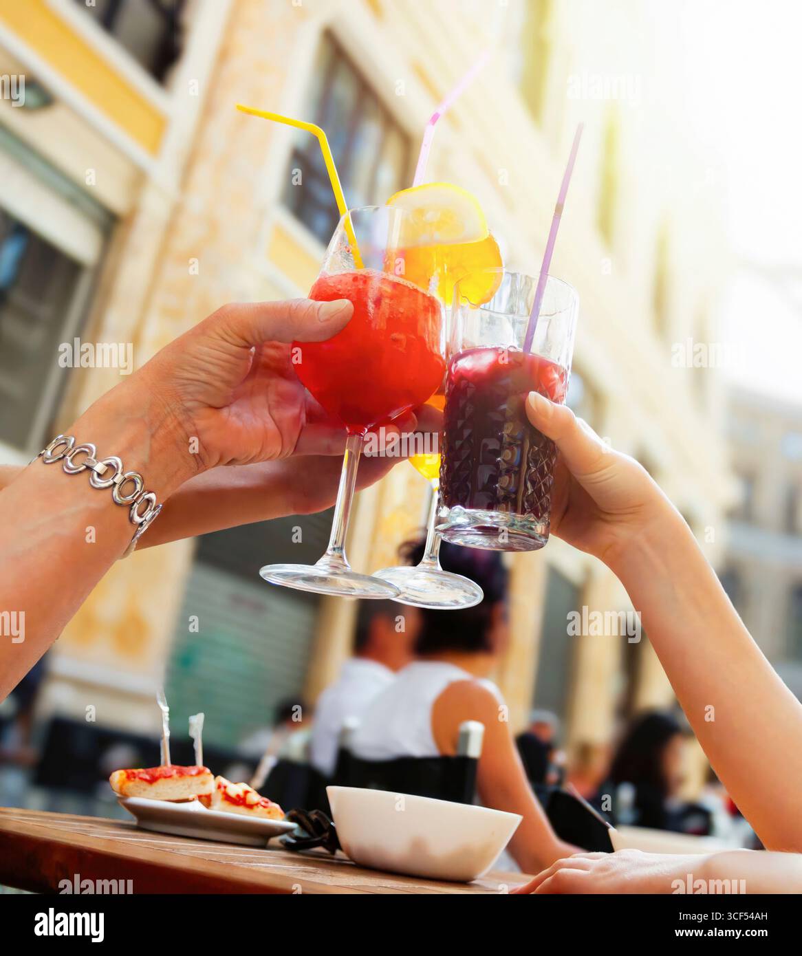 Eine Gruppe von Freunden, die an einem Restauranttisch in Italien bunte Cocktails mit Strohhalmen anstoßen und an einem sonnigen Tag die Happy Hour mit Vorspeisen und Getränken genießen Stockfoto