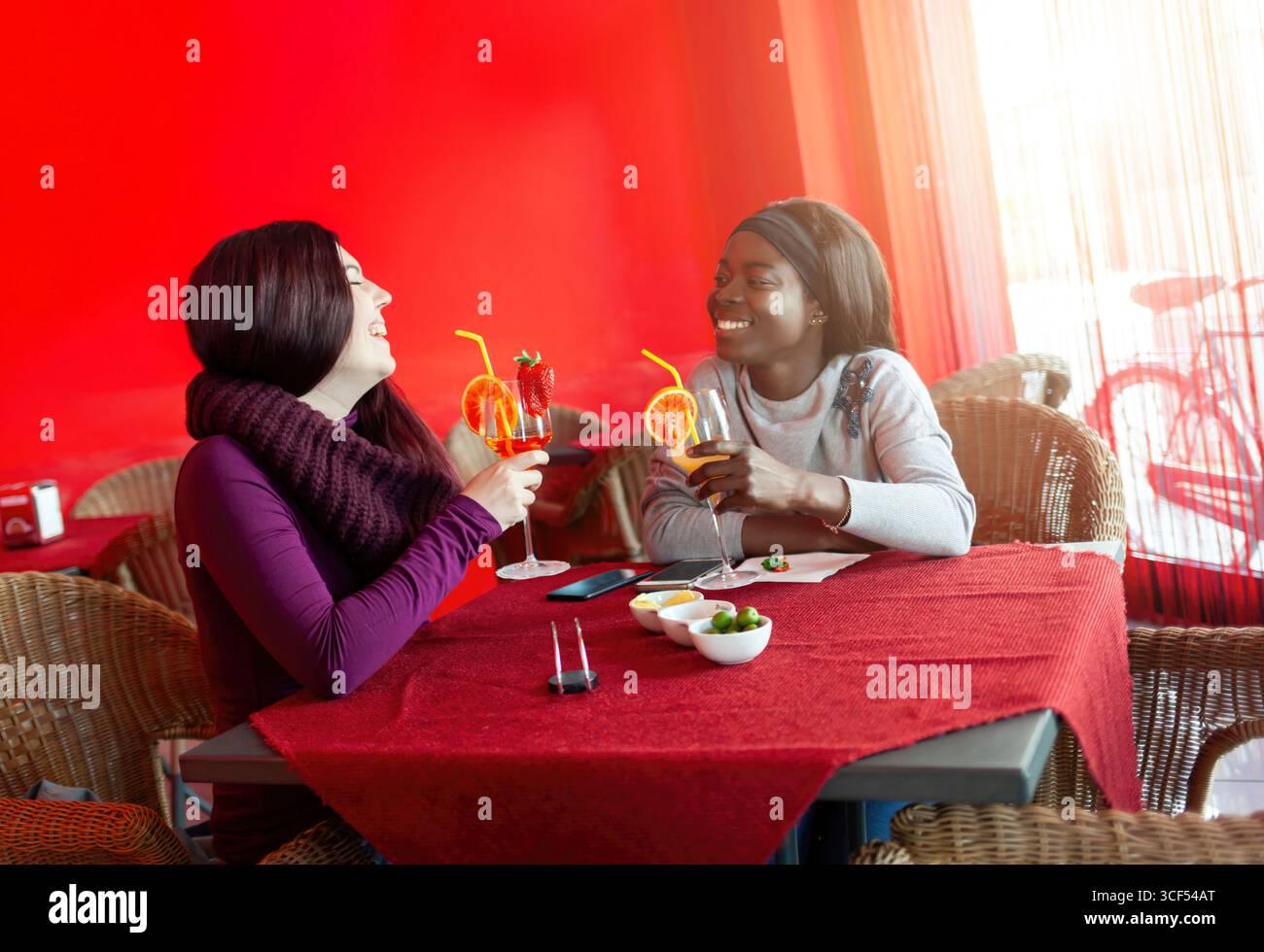 Zwei ethnische Freundinnen genießen Cocktails und Appetithäppchen an einer Bar, lachen und unterhalten sich in einer lebhaften roten Atmosphäre und feiern Freundschaft und Zweisamkeit Stockfoto