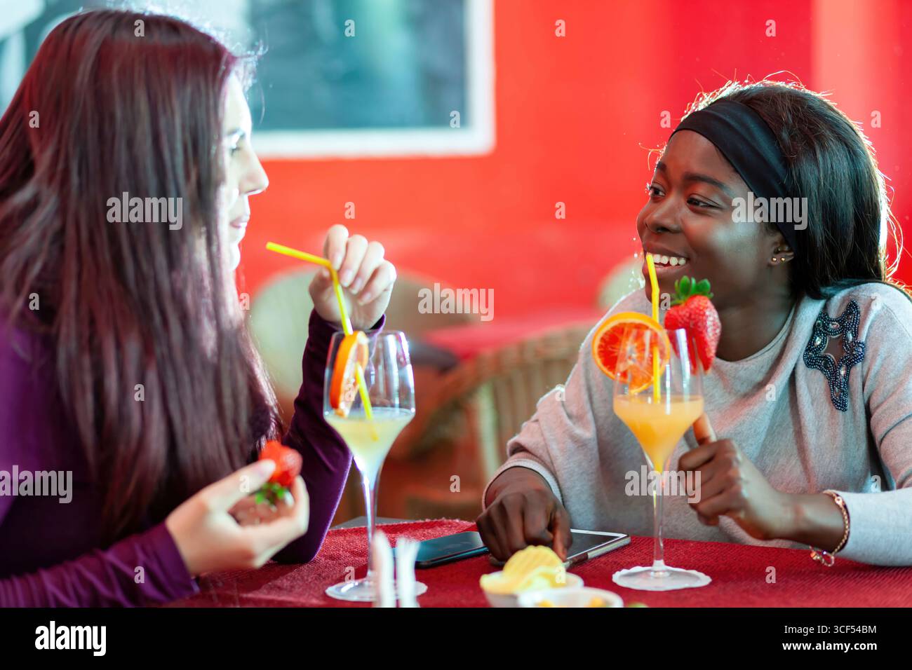Zwei Frauen sitzen an einem Tisch in einer Bar, trinken Cocktails, die mit Fruchtscheiben und Erdbeeren dekoriert sind, und führen ein angenehmes Gespräch Stockfoto
