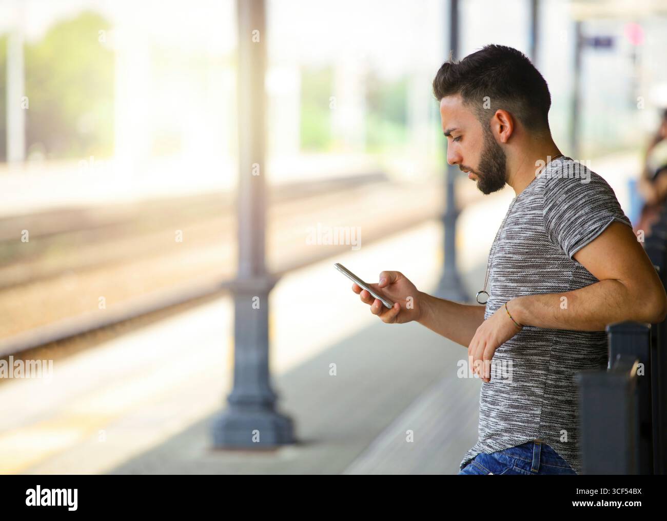 Lässig gekleideter junger Mann, der sich an einem Geländer lehnt und sein Smartphone benutzt, um in Verbindung zu bleiben, während er an einem hellen, sonnigen Tag auf einen Zug am Bahnhof wartet Stockfoto