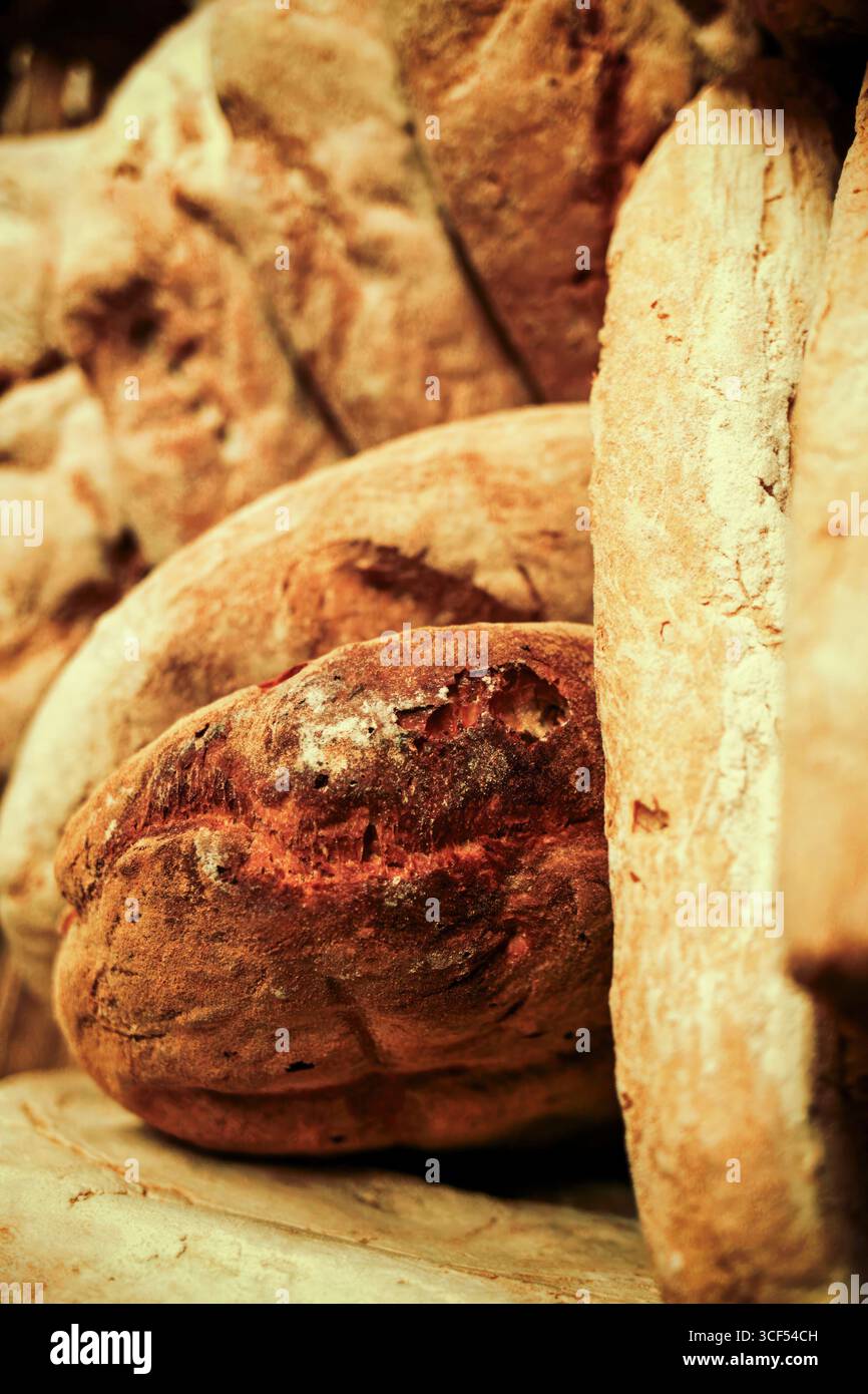 Nahaufnahme verschiedener Arten frisch gebackener Brotlaibe mit einer Vielzahl von Texturen und Krusten, die auf einem Holzregal in einer Bäckerei ruhen, was eine warme und einladende Atmosphäre suggeriert Stockfoto