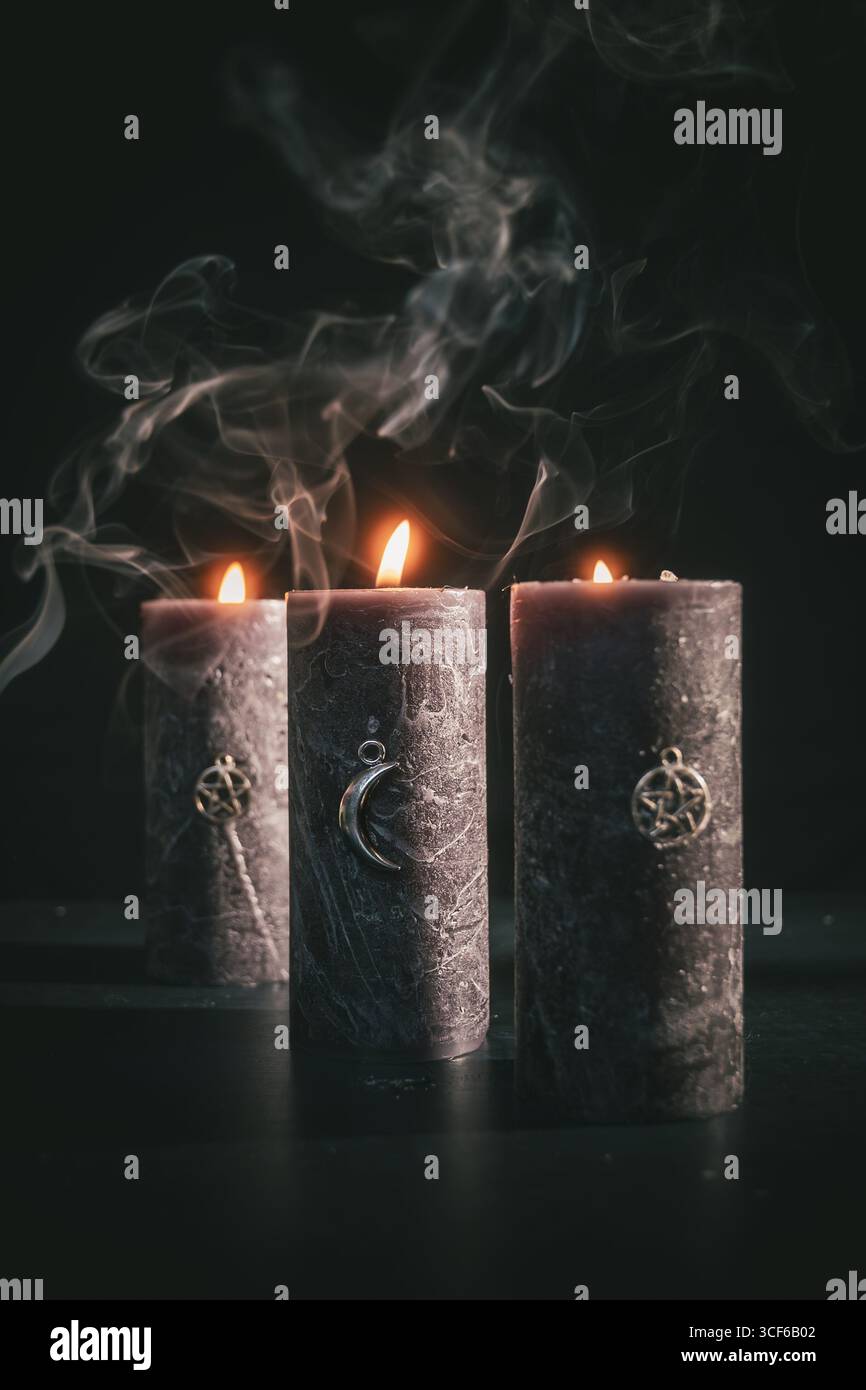 Drei schwarze Kerzen mit glühenden Flammen und okkulten Anhängern, umgeben von aufsteigendem Rauch in einer dunklen, mystischen Atmosphäre. Stockfoto