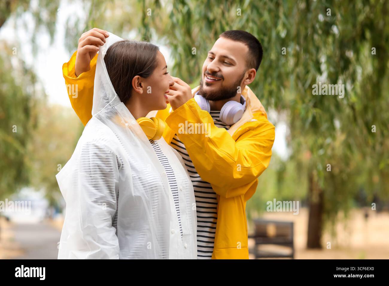 Junger Mann, der den Kopf seiner Freundin mit Regenmantel im Park bedeckt Stockfoto