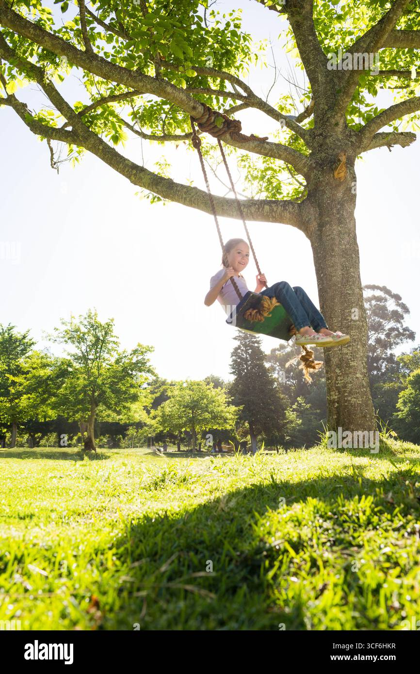 Kindermädchen schwingend auf Seilschaukel unter Laubbäumen im Park mit Holzsitz, Kopierraum Stockfoto