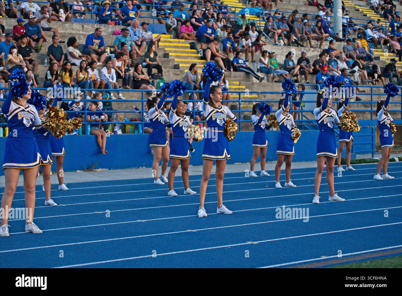 Cheerleader in leuchtenden blauen, gelben und weißen Uniformen schlagen dynamische Posen unter dem goldenen Sonnenuntergangshimmel von Hawai'i und strahlen Stolz, Geist und Einheit aus. Stockfoto