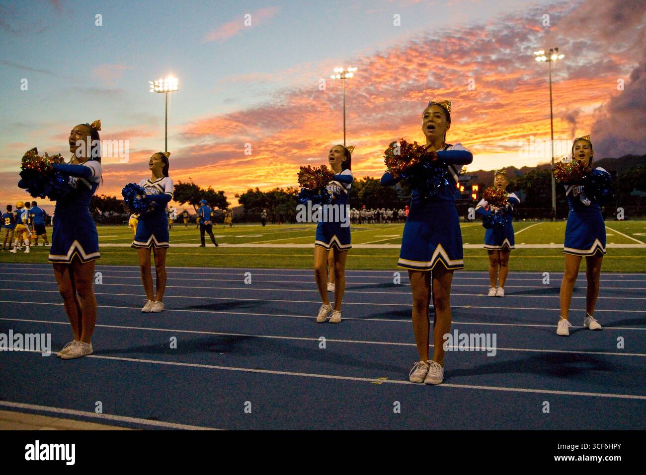 Cheerleader in leuchtenden blauen, gelben und weißen Uniformen schlagen dynamische Posen unter dem goldenen Sonnenuntergangshimmel von Hawai'i und strahlen Stolz, Geist und Einheit aus. Stockfoto